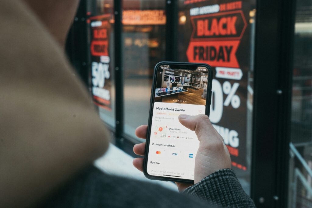 personne de dos tenant un smartphone affichant une page d'une boutique en ligne, vitrine d'un magasin en arrière-plan avec des stickers promotionnels sur le Black Friday - AVICOM'